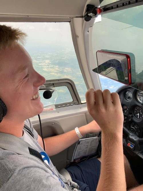 A person wearing a headset smiles while piloting a small airplane. They hold a control yoke and have a paper checklist on their lap. The cockpit window shows a view of the sky and landscape below.