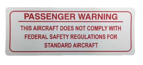 Image of a warning sign reading: "Passenger Warning - This aircraft does not comply with federal safety regulations for standard aircraft." The text is in red on a white background, emphasizing safety concerns.