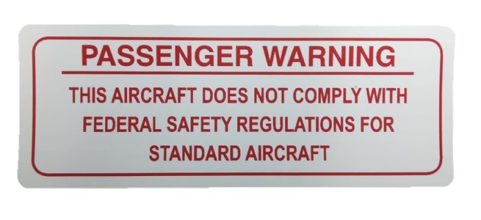 Image of a warning sign reading: "Passenger Warning - This aircraft does not comply with federal safety regulations for standard aircraft." The text is in red on a white background, emphasizing safety concerns.