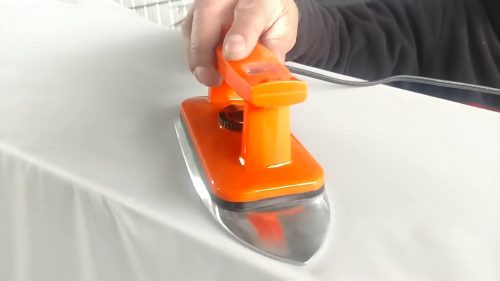 A person uses the Fabric Pro Iron with an orange handle to press a white fabric on an ironing board.