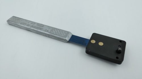 A Plate Nut Drill Jig with a long metal handle and a black rectangular base featuring two gold-colored circles and a small black knob, set against a plain white background.