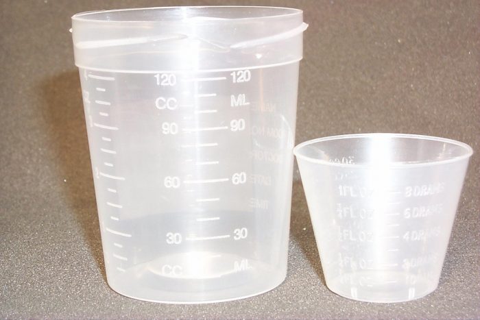Two clear plastic measuring cups are shown. The larger cup has measurement markings up to 120 milliliters, and the smaller is labeled as a 1 oz. plastic measuring mixing cup. Both are on a textured surface.