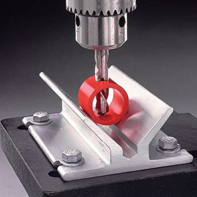 A drill is positioned above a red cylindrical object held in place by a CENTER-IT HOLE V-BLOCK. The V-block is mounted on a black base, with bolts securing it.