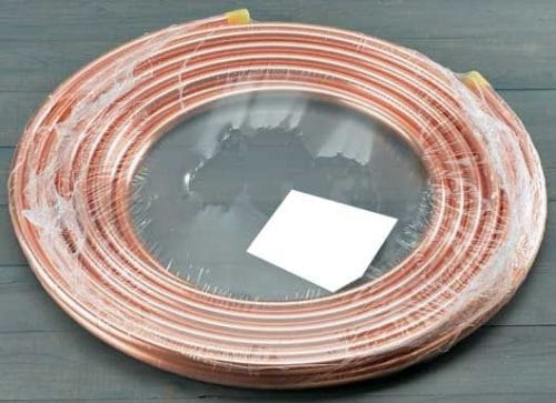 A coiled copper pipe wrapped in clear plastic sits on a gray surface. A small white rectangular label is placed on top of the coil.