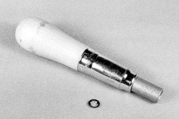 A metal and wood dental tool with a cylindrical handle and a smaller circular tip is placed on a flat surface. A small circular washer lies nearby.