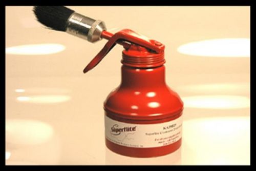 Red oil can with a brush attached to the spout, placed on a reflective surface. The can has a label with text and a logo.