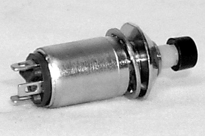 A small metallic push button switch with a cylindrical body, two metal prongs for connections, and a black button on one end, lying on a plain surface.