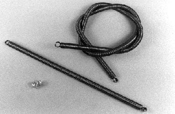 A coiled metal cable with looped ends is positioned above a straight metal rod and two small spherical objects on a plain surface.