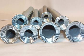 Several metal pipes of varying diameters are arranged horizontally on a plain surface, with the open ends facing the viewer. The pipes have a shiny, reflective surface and concentric grooves at the openings.