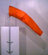 Orange windsock mounted on a metal pole, slightly inflated, indicating wind direction and strength. The background is a plain, light-colored wall.